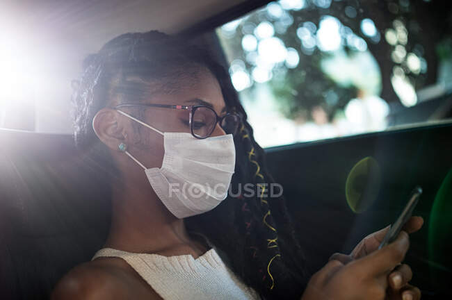 Young afro latin woman in face mask uses smartphone in the back seat of a car — Stock Photo
