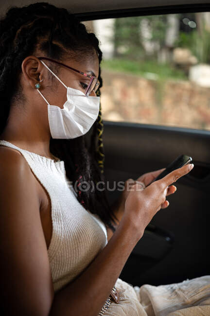 Молода африкано латиноамериканська жінка в масці використовує смартфон на задньому сидінні автомобіля. — стокове фото