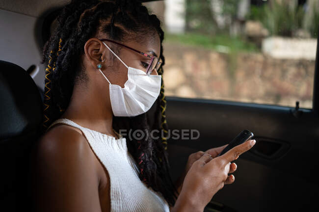 Young afro latin woman in face mask uses smartphone in the back seat of a car — Stock Photo