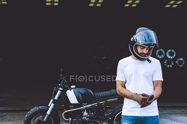 Corpo inteiro de jovem motociclista masculino étnico grave em roupas casuais e boné de pé perto de moto perto de guindastes industriais à beira-mar — Fotografia de Stock