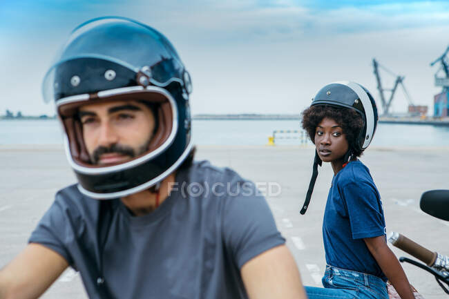 Elegante joven pareja multiétnica en traje casual y cascos descansando en el paseo marítimo después de montar en motocicleta - foto de stock