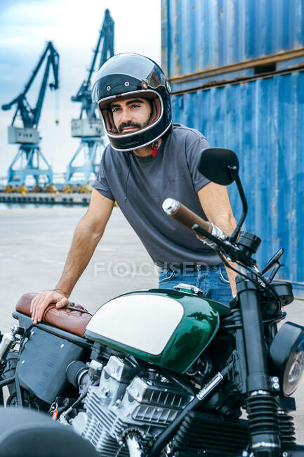 Auto asegurado joven hombre barbudo étnico en ropa elegante y casco apoyado en la motocicleta moderna y mirando hacia otro lado en la zona industrial - foto de stock