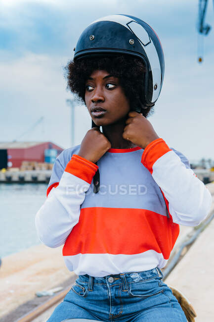 Concentrated young black female biker with Afro hair in trendy outfit and helmet while sitting on motorcycle at seaside — Stock Photo