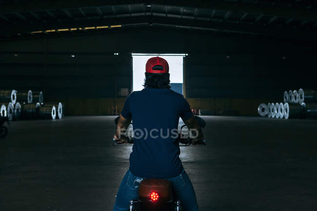 Vista trasera de ciclista masculino irreconocible en ropa casual y gorra montando motocicleta moderna en amplio hangar industrial con pila de bobinas de chapa metálica - foto de stock