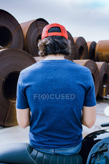 Back view of unrecognizable male with dark hair in casual outfit and cap standing near stack of metal sheet coils in industrial factory — Stock Photo