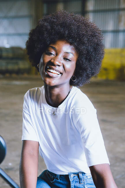 Cheerful young African American lady with curly hair in white t shirt and jeans smiling and looking at camera in industrial area — Stock Photo