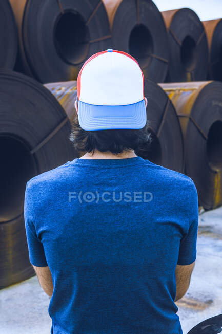 Back view of unrecognizable male with dark hair in casual outfit and cap standing near stack of metal sheet coils in industrial factory — Stock Photo
