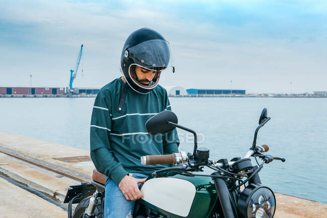Motociclista masculino con traje casual y casco protector sentado en motocicleta en terraplén cerca del mar en el sitio de construcción - foto de stock