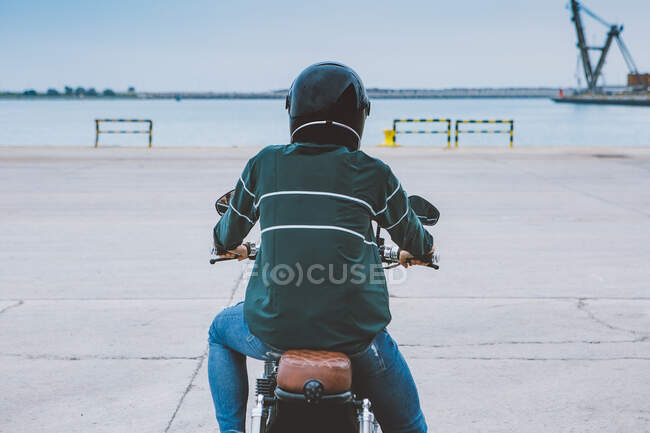 Vista trasera de ciclista masculino irreconocible en traje casual y casco de protección sentado en motocicleta en terraplén cerca del mar en el sitio de construcción - foto de stock
