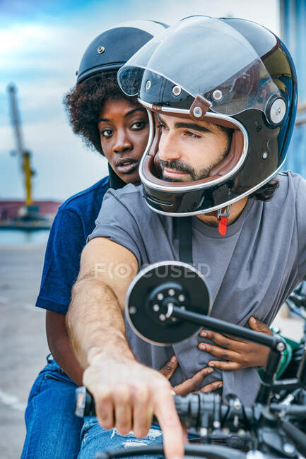 Stylish young ethnic guy and African American lady in casual outfits and helmets sitting on motorcycle on city street — Stock Photo