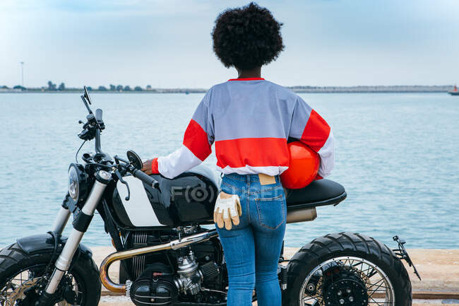Back view of unrecognizable young African American lady with curly hair in stylish outfit standing near modern motorbike with helmet in hand and admiring sea — Stock Photo