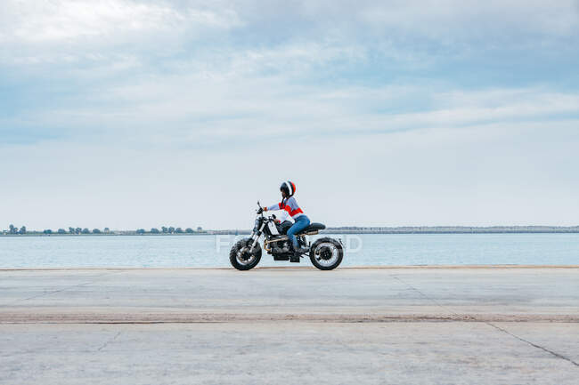 Vista lateral do motociclista feminino irreconhecível em roupas elegantes e motocicleta de equitação de capacete na estrada de asfalto vazia perto do mar contra o céu azul nublado — Fotografia de Stock