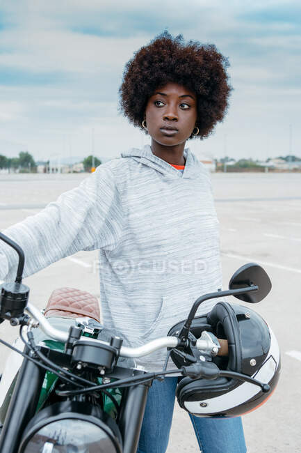 Self assured young African American woman with curly hair in stylish outfit leaning on motorbike and looking away in city outskirts — Stock Photo