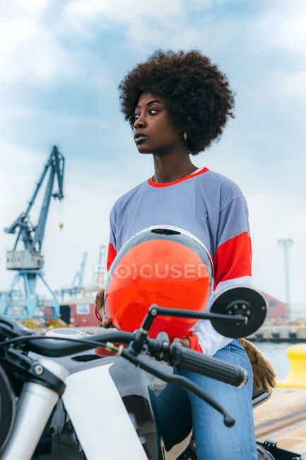 Joven motorista negra concentrada con pelo afro en traje de moda y casco mientras está sentada en motocicleta en la playa - foto de stock