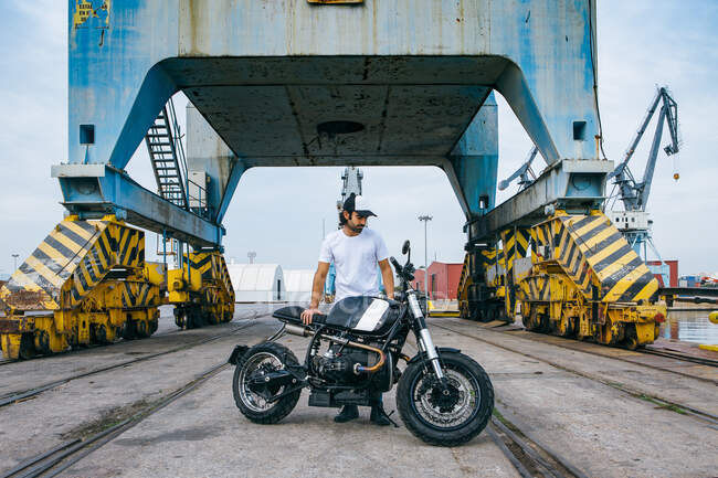 Corpo inteiro de jovem motociclista masculino étnico grave em roupas casuais e boné de pé perto de moto perto de guindastes industriais à beira-mar — Fotografia de Stock