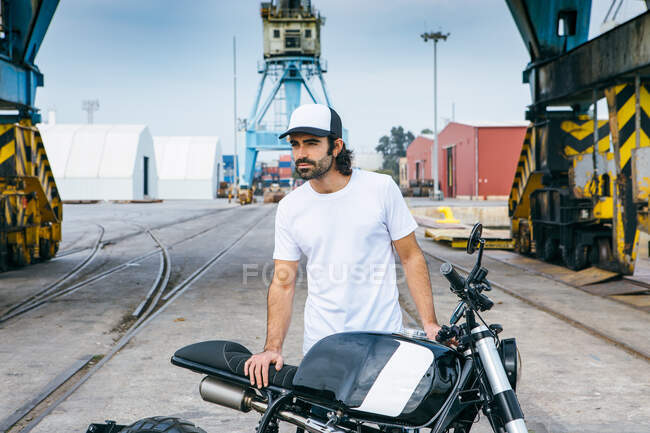 Vista lateral do motociclista masculino irreconhecível em roupa casual e capacete de proteção sentado na motocicleta em aterro perto do mar no local de construção — Fotografia de Stock
