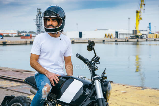 Confident young bearded ethnic guy in white t shirt and jeans while standing motorcycle at seaside — Stock Photo