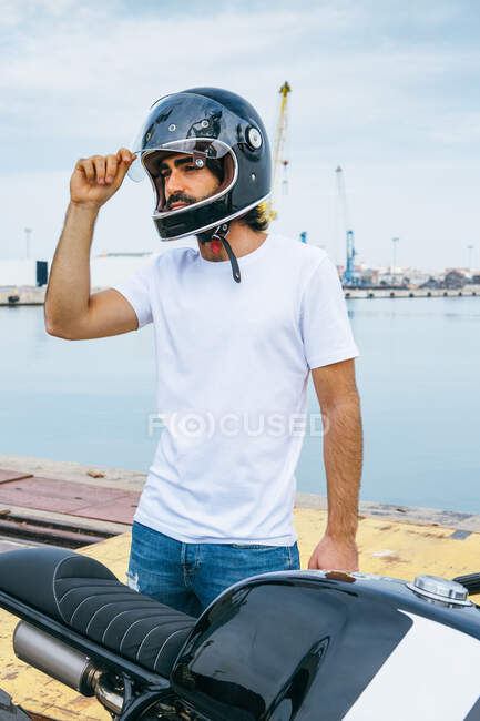 Confident young bearded ethnic guy in white t shirt and jeans tying helmet while standing motorcycle at seaside — Stock Photo