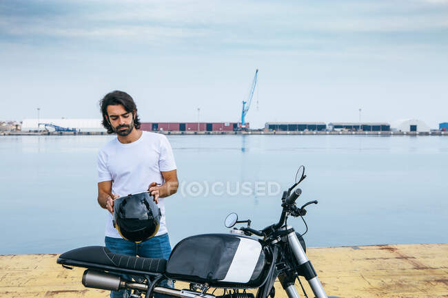 Confident young bearded ethnic guy in white t shirt and jeans while standing motorcycle at seaside — Stock Photo