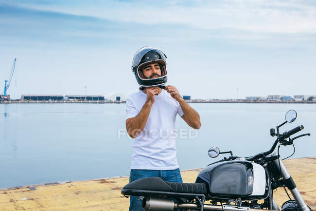 Уверенный молодой бородатый этнический парень в белой футболке и джинсах, завязывающий шлем, стоя на мотоцикле у моря — стоковое фото
