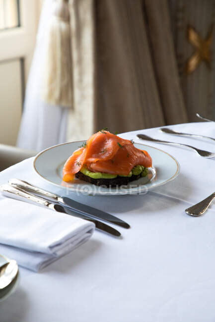 Высокий угол тарелки с вкусными тостами авокадо с яичницей-пашот и копченым лососем подается на столе со столовыми приборами и кофейной чашкой во время завтрака в ресторане отеля — стоковое фото