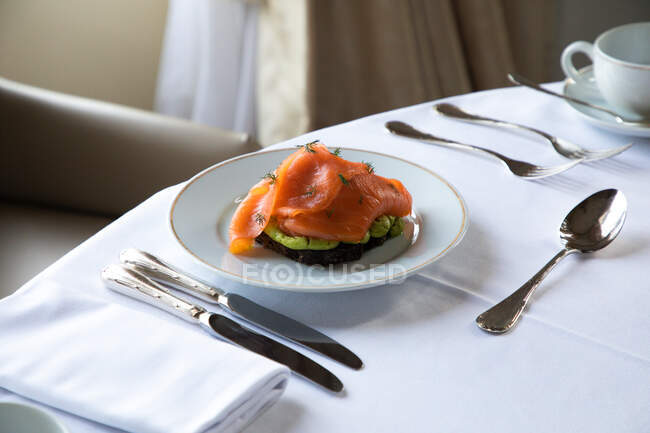 Alto ángulo de plato con deliciosas tostadas de aguacate con huevo escalfado y salmón ahumado servido en la mesa con cubiertos y taza de café durante el desayuno en el restaurante del hotel - foto de stock
