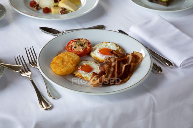 Alto ángulo de apetitosos huevos fritos con rodajas de tocino servidas en plato con tomate relleno y mozzarella crujiente y colocadas en la mesa con cutler y taza de café durante el desayuno - foto de stock