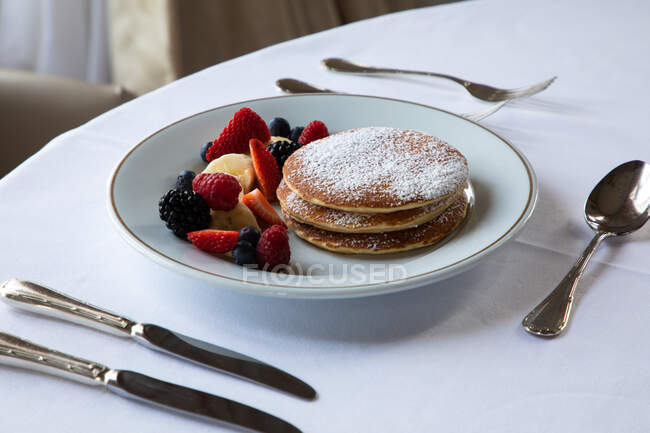 From above plate of tasty pancakes with sugar powder served with assorted healthy berries and banana slices during breakfast in cafe — Stock Photo