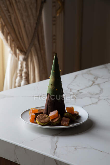 Шоколадный десерт в форме ёлки на тарелке с различными печеньями и мармеладом, подаваемый на мраморном столе в стильном ресторане — стоковое фото
