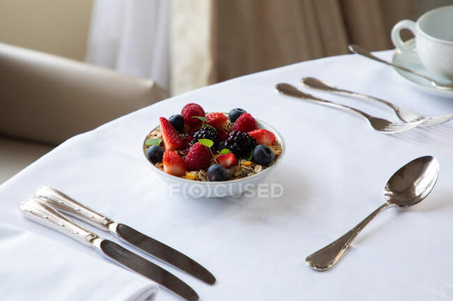 З верхньої миски здорового муеслі, увінчаного свіжими збитими ягодами, подається на білому столі зі сріблястим посудом і чашечкою кави під час сніданку. — стокове фото
