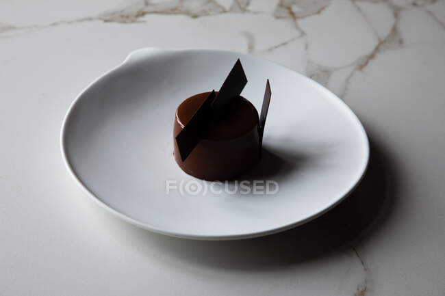 Високий кут смачного шоколадного глазурованого торта, прикрашеного їстівними прикрасами, що подаються на білій тарілці і поміщені на мармуровий стіл — стокове фото