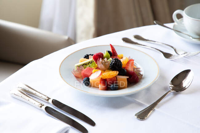 From above of tasty fruit salad with assorted berries kiwi and watermelon served on white plate on table with cup of coffee and cutlery in hotel restaurant — Stock Photo