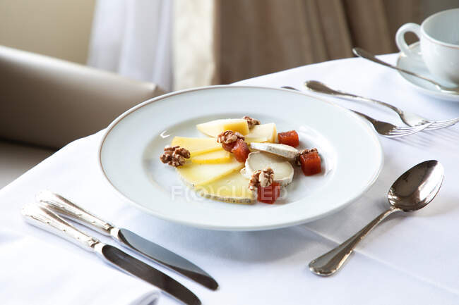 Сверху разнообразные ломтики сыра в тарелке подаются с грецкими орехами и кубиками копченого лосося на белом столе возле кофейной чашки и столовых приборов — стоковое фото