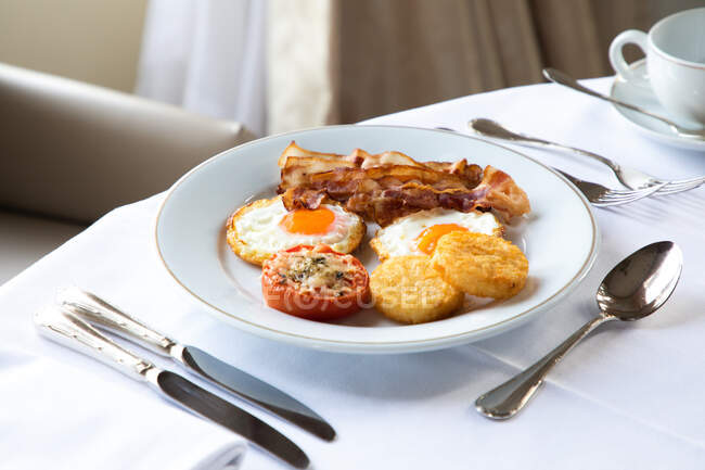 High angle of appetizing fried eggs with bacon slices served on plate with stuffed tomato and crispy mozzarella and placed on table with cutler and cup of coffee during breakfast — Stock Photo