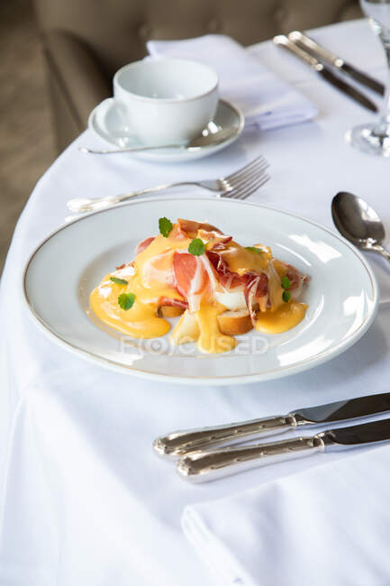 From above of appetizing Eggs Benedict dish with bacon topped with hollandaise sauce served in white plate on table with cup of coffee and silverware in restaurant during brunch — Stock Photo