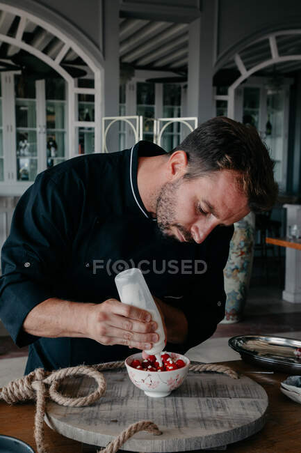 Concentrado jovem chef masculino em uniforme usando seringa creme ao decorar tigela com bagas frescas em pé à mesa em restaurante elegante — Fotografia de Stock