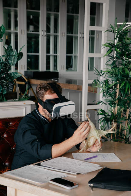 Chef masculino irreconhecível em uniforme sentado à mesa com vários documentos e smartphones e segurando milho grelhado enquanto experimenta a realidade virtual no fone de ouvido VR no restaurante — Fotografia de Stock