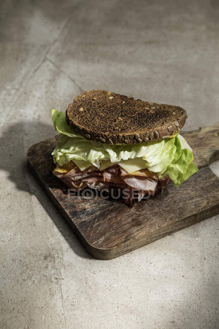 Высокий угол аппетитного сэндвича со свежим корневым хлебом над листьями салата и беконом на деревянной доске — стоковое фото