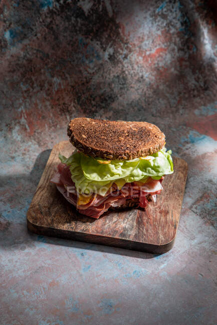 Alto ángulo de bocadillo apetitoso con pan crujiente fresco sobre hojas de lechuga y tocino sobre tabla de madera - foto de stock