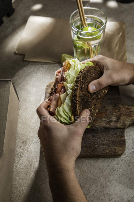 Зверху врожаю анонімний клієнт ресторану, який їсть смачний бутерброд з тостами бекону та листям салату з водою з лаймом — стокове фото
