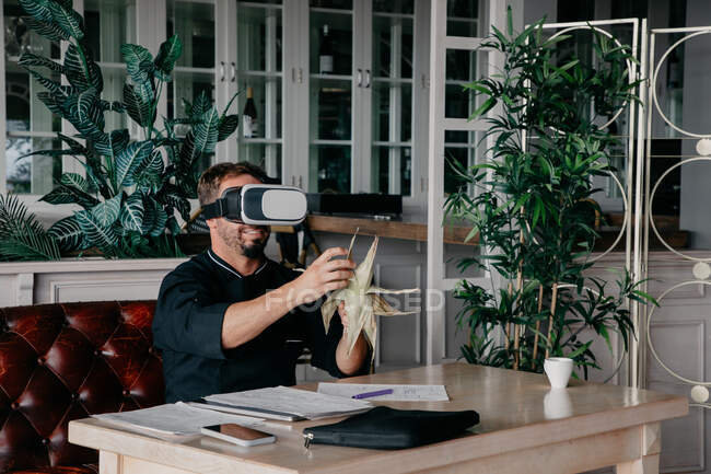Unrecognizable male chef in uniform sitting at table with various documents and smartphone and holding grilled corn while experiencing virtual reality in VR headset in restaurant — Stock Photo