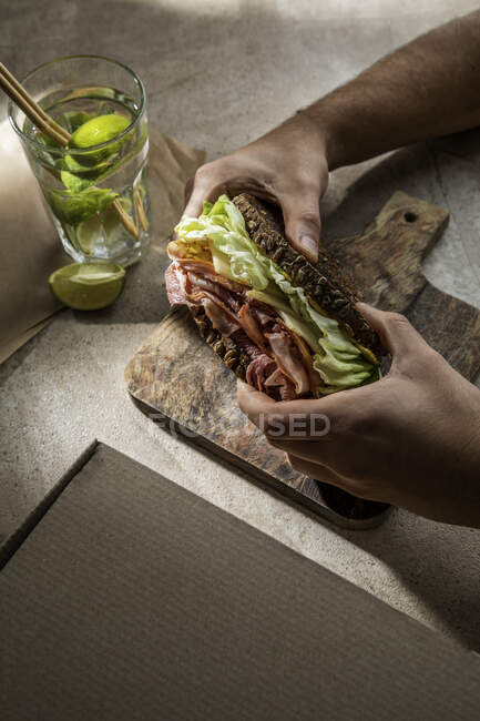 Сверху урожая анонимный клиент ресторана ест вкусный сэндвич из тостов с беконом и листья салата с водой с лаймом — стоковое фото