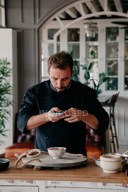 Konzentrierter männlicher Koch in Uniform fotografiert mit dem Handy die Mahlzeit in der Schüssel im Restaurant — Stockfoto