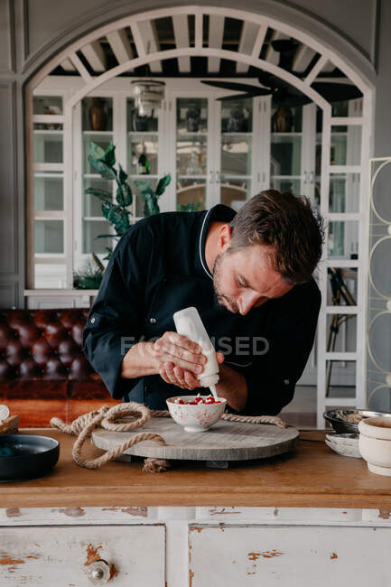 Concentrated young male chef in uniform using cream syringe while decorating bowl with fresh berries standing at table in stylish restaurant — Stock Photo