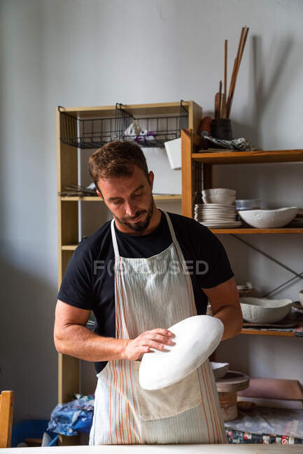 Concentrated young bearded male potter in casual clothes and apron creating white ceramic plate while working in studio — Stock Photo