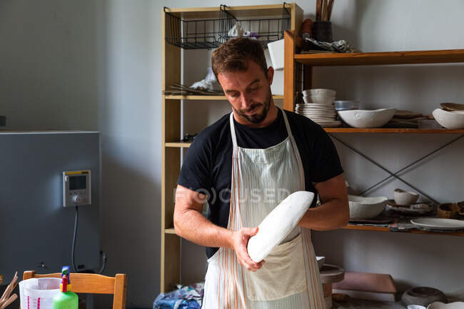 Alfarero masculino barbudo joven concentrado en ropa casual y delantal creando placa de cerámica blanca mientras trabaja en el estudio - foto de stock
