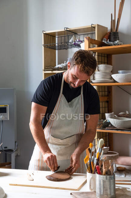 Позитивный молодой мужчина-мастер в повседневной одежде и фартуке улыбается, создавая глиняную посуду, стоя за столом в мастерской — стоковое фото