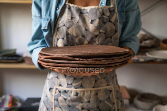 Crop unrecognizable female artisan in casual outfit and apron holding stack of flat clay plates while working in art studio — Stock Photo