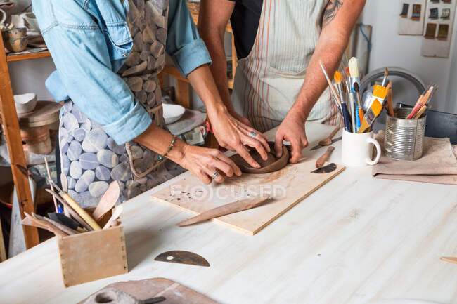 Cropped unrecognizable female and male in casual clothes and aprons smiling while creating clay plate during pottery class in workshop — Stock Photo