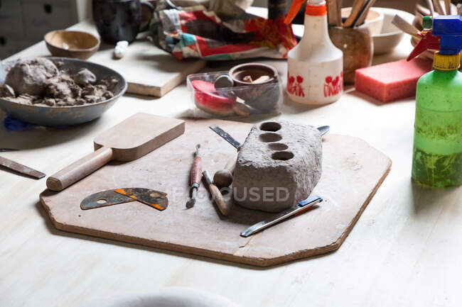 De dessus morceau d'argile placé sur une planche de bois avec divers outils de sculpture et nervure de poterie métallique en atelier — Photo de stock